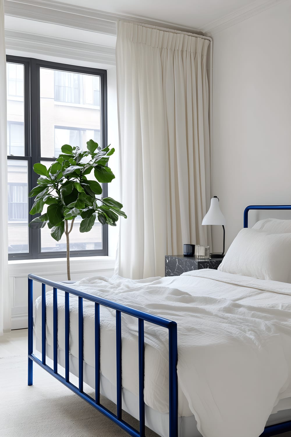 A minimalist bedroom featuring a bed with cobalt blue metal frames and bedside tables, adorned with white linen. There's a matte black marble nightstand with a minimalist lamp on one side. Large windows with sheer white curtains allow ample natural light to fill the room, highlighting a single emerald green potted plant.