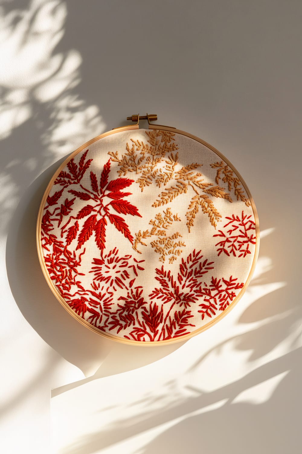 A single embroidery hoop featuring intricate red and gold leaf motifs hangs on a minimalist white wall. Gentle shadows enhance the detailed stitching of the designs, creating a festive and artistic display.