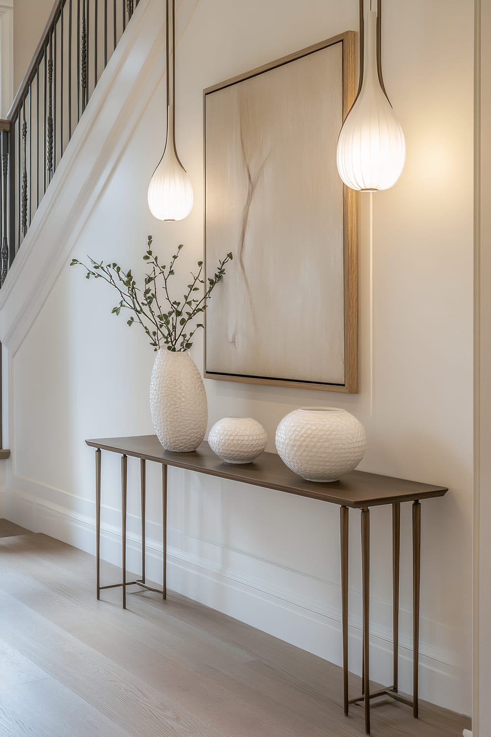 A stylish entryway featuring a bronze-toned console table adorned with three white ceramic vases. The larger vase holds green sprigs, while the two smaller spherical vases are empty. Two overhead pendant lights with white, tear-shaped shades softly illuminate the space. A large abstract painting with neutral tones hangs centrally on the wall above the console. The staircase on the left has black metal balusters and a wooden railing. The floor is a light wood.