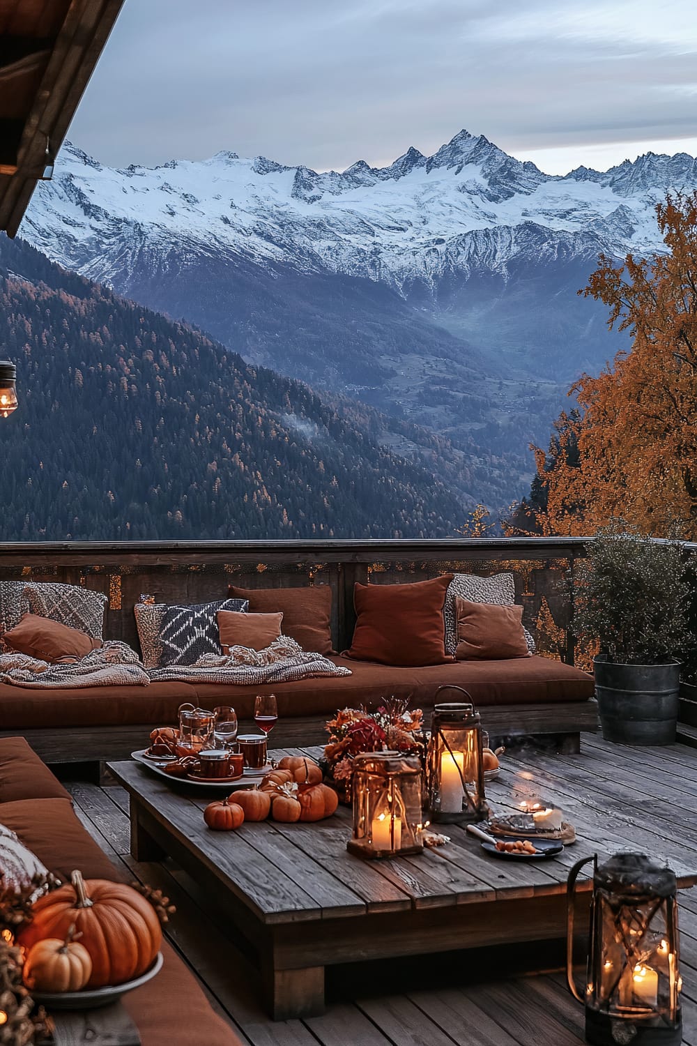 A serene outdoor seating area with a panoramic view of snow-capped mountains and autumnal forests. The seating area has rust-colored cushions and various patterned pillows on a wooden bench. A rustic wooden coffee table is adorned with small pumpkins, candles, and drinks. A potted plant sits in the corner, and the setting is illuminated by candlelight and a hanging lantern.