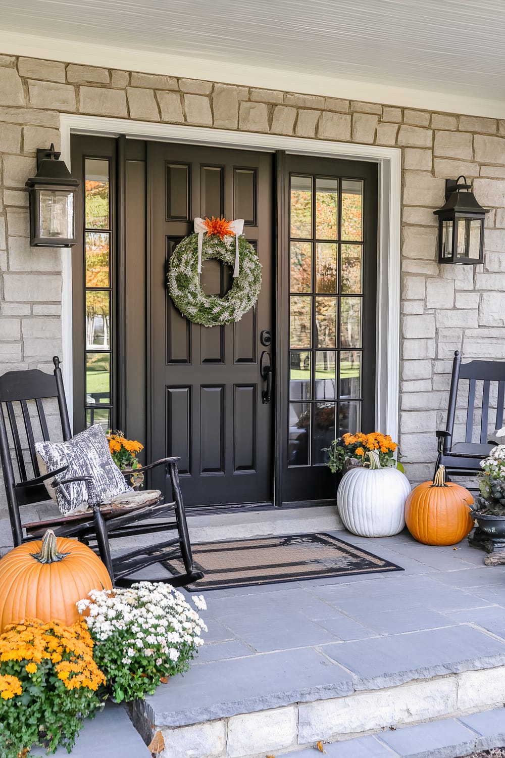 An inviting front porch decorated for autumn. The door is black with glass side panels, adorned with a wreath made of greenery and a small orange bow. On either side are two black rocking chairs, one holding a cushion with a monochromatic design. Various pumpkins in orange and white, along with pots of yellow mums, decorate the steps and floor of the porch. Large black lanterns are fixed to the stone walls beside the door.