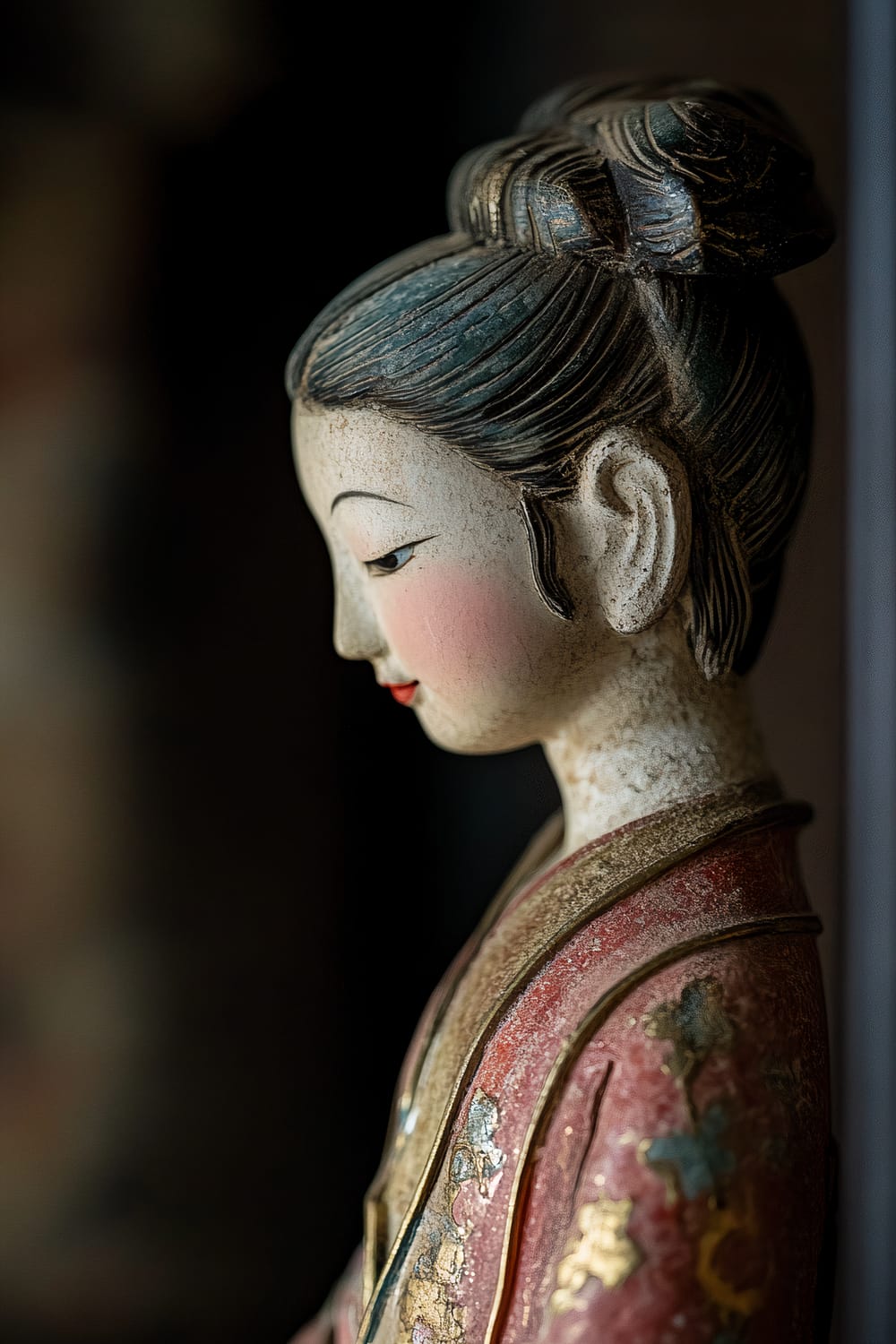 A close-up image of a handcrafted antique East-Asian figurine seen in profile. The figurine, painted with vibrant colors of pink, gold, and blue, showcases elegant attire and a serene expression. The figurine is displayed on a dark wood shelf against a dark, blurred background, highlighted by soft ambient lighting.