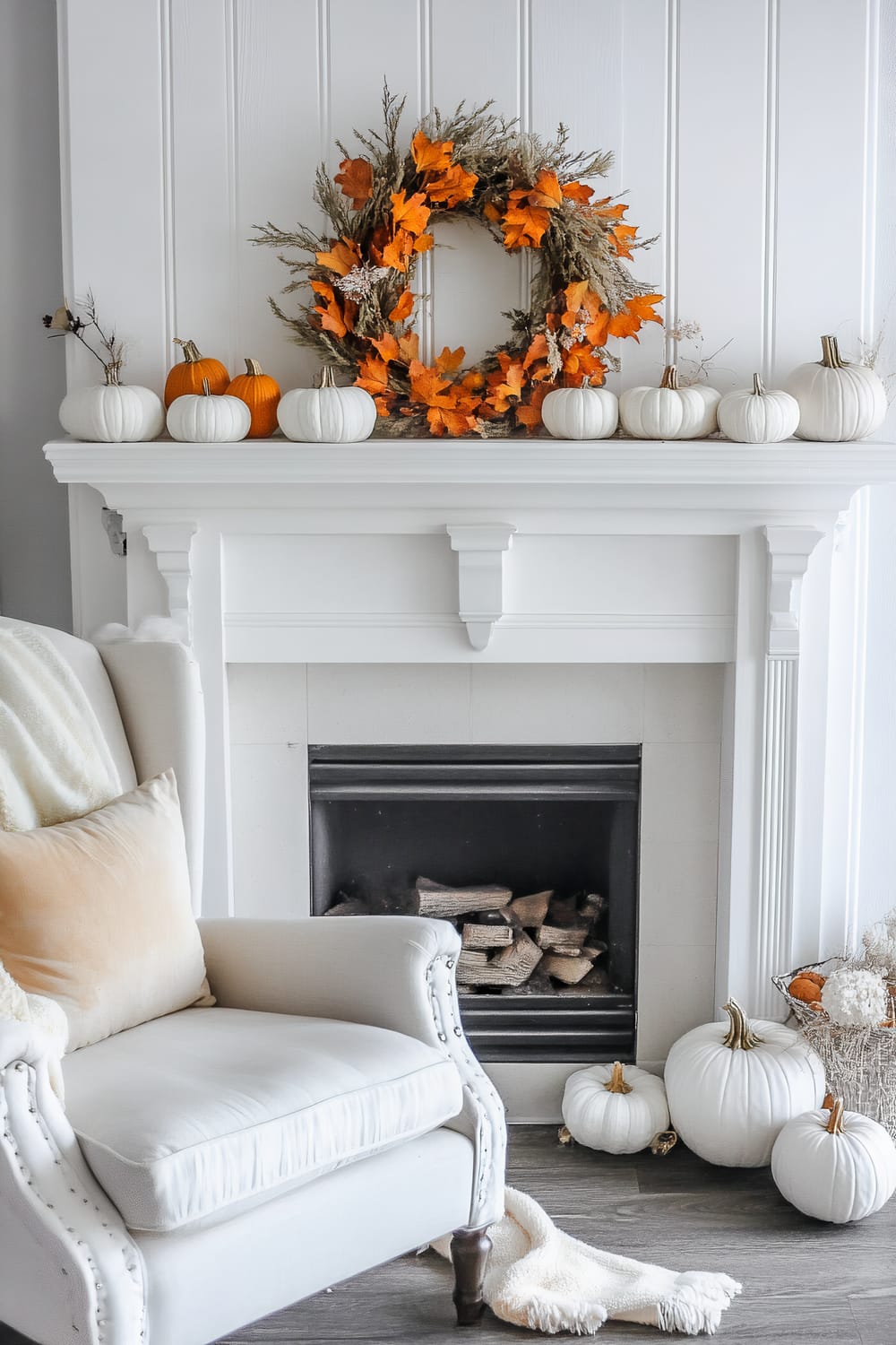 A sophisticated interior scene showcasing a white fireplace mantle adorned with an autumn wreath made of orange leaves and dried foliage. Surrounding the wreath are white and orange pumpkins arranged in a symmetrical pattern. Below the mantle lies a stack of firewood inside the black fireplace. To the left is an elegant white armchair with beige cushions and a white throw blanket draped over it. More white pumpkins are placed on the floor to the right of the fireplace, next to a basket filled with warm-toned autumn decor.