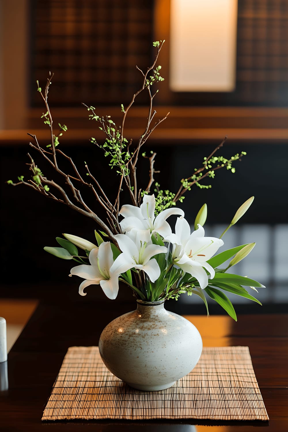 A traditional, yet refined Ikebana-inspired centerpiece on a dark wooden table. The centerpiece features minimalist branches, white lilies, and subtle green leaves in a low ceramic vase. The staging includes a simple tatami mat runner underneath. Accents around the table are smooth river stones, with soft ambient lighting providing a tranquil mood.