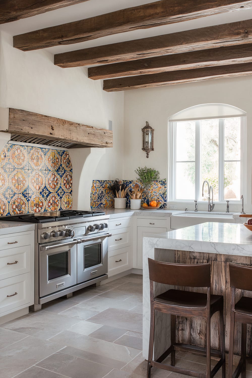 A rustic kitchen showcases a blend of earthy and elegant elements. Exposed wooden beams accentuate the white ceiling and walls, while the focal point is a kitchen backsplash with intricate, colorful tiles in Mediterranean designs. A large stainless steel double oven and range with six burners sits beneath a substantial wooden range hood. The marble countertops contrast with the wooden island, creating a balance. White cabinetry with dark metal handles adds a touch of minimalism. Large wooden barstools at the island complement the wooden beams. A single lantern-style wall sconce and an arched window provide natural light and an outdoor view, with oranges and a potted plant adding a pop of color on the counter.