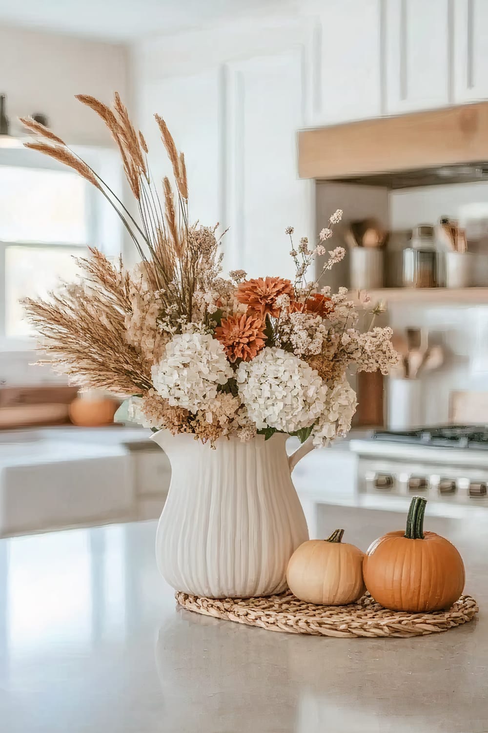 A white ceramic vase filled with a bouquet of dried and fresh flowers, including white hydrangeas and orange chrysanthemums, standing on a white kitchen counter. Next to the vase, there are two small pumpkins, one orange and one pale yellow, placed on a woven placemat. The kitchen in the background features white cabinetry and wooden shelves with various kitchen items.