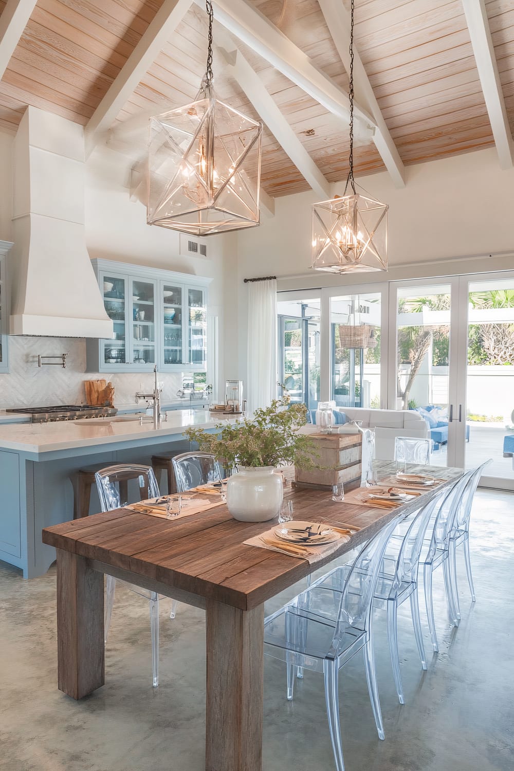 A sophisticated dining and kitchen space featuring a large wooden dining table with clear, ghost-style chairs. The table is set with neutral place settings and a white vase with greenery as a centerpiece. Above the table hang two geometric pendant lights with exposed bulbs. The background includes a kitchen with light blue cabinetry, a white range hood, and a marble countertop. Large glass doors open to an outdoor patio area, bringing natural light into the space. The ceiling is vaulted with exposed wooden beams.