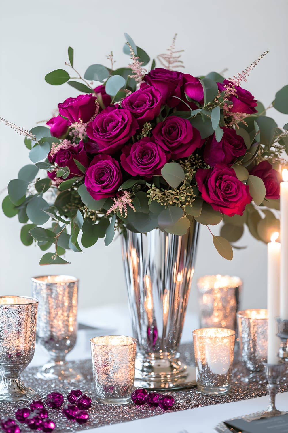 A modern centerpiece display featuring a tall silver mirrored vase holding vibrant fuchsia roses and eucalyptus branches. The vase is surrounded by slender silver candle holders with white candles and scattered fuchsia glass gems. All these accessories are presented on a white table with a sparkling silver sequin runner under cool, focused lighting.