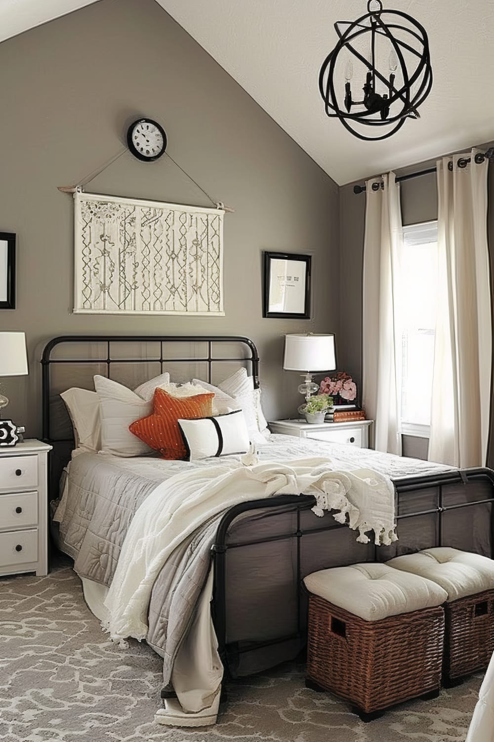 A modern, well-decorated bedroom with a high vaulted ceiling, featuring a neutral color palette. The bed has an iron frame and is adorned with white, grey, and orange pillows, along with a white throw blanket. Two nightstands with lamps flank the bed, and a piece of macramé art hangs above the headboard, beneath a black wall clock. The room is illuminated by natural light coming through a window with long, white curtains. A contemporary chandelier with a spherical design hangs from the ceiling. Below the bed, two woven storage baskets with cushioned tops are set against a patterned area rug.