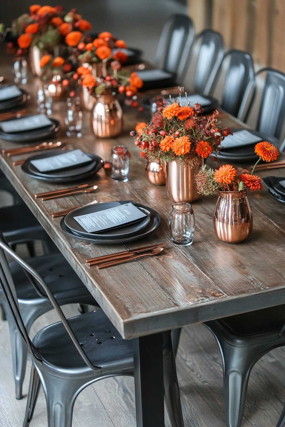 An elegantly set dining table with a rustic wooden surface, decorated with bright orange flowers in copper vases and minimalist black plates with menus placed on top. Copper cutlery and clear glasses accompany the place settings, while metal chairs surround the table.