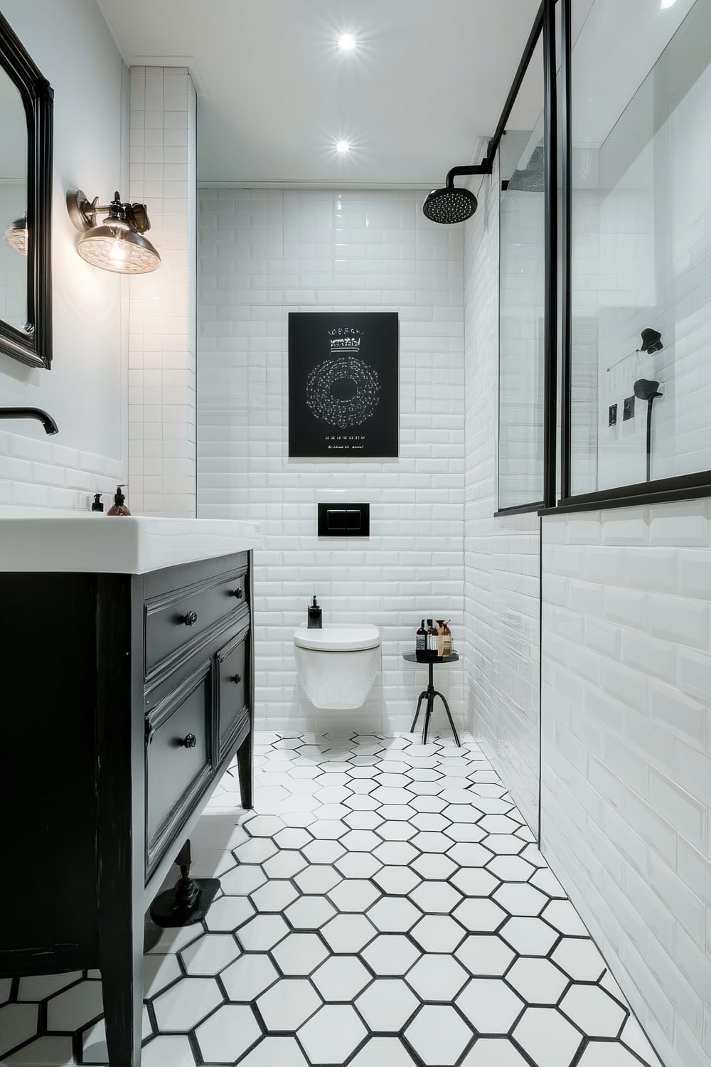 A modern bathroom featuring white subway tile walls and hexagonal tile flooring. The bathroom includes a black-framed walk-in shower with a rainfall showerhead and glass doors. A black vanity with a white countertop is on the left side, complemented by an industrial-style wall sconce with an Edison bulb. In the center, a wall-mounted toilet is positioned beneath a black-framed piece of art. Beside the toilet, there is a small black side table with various toiletries.