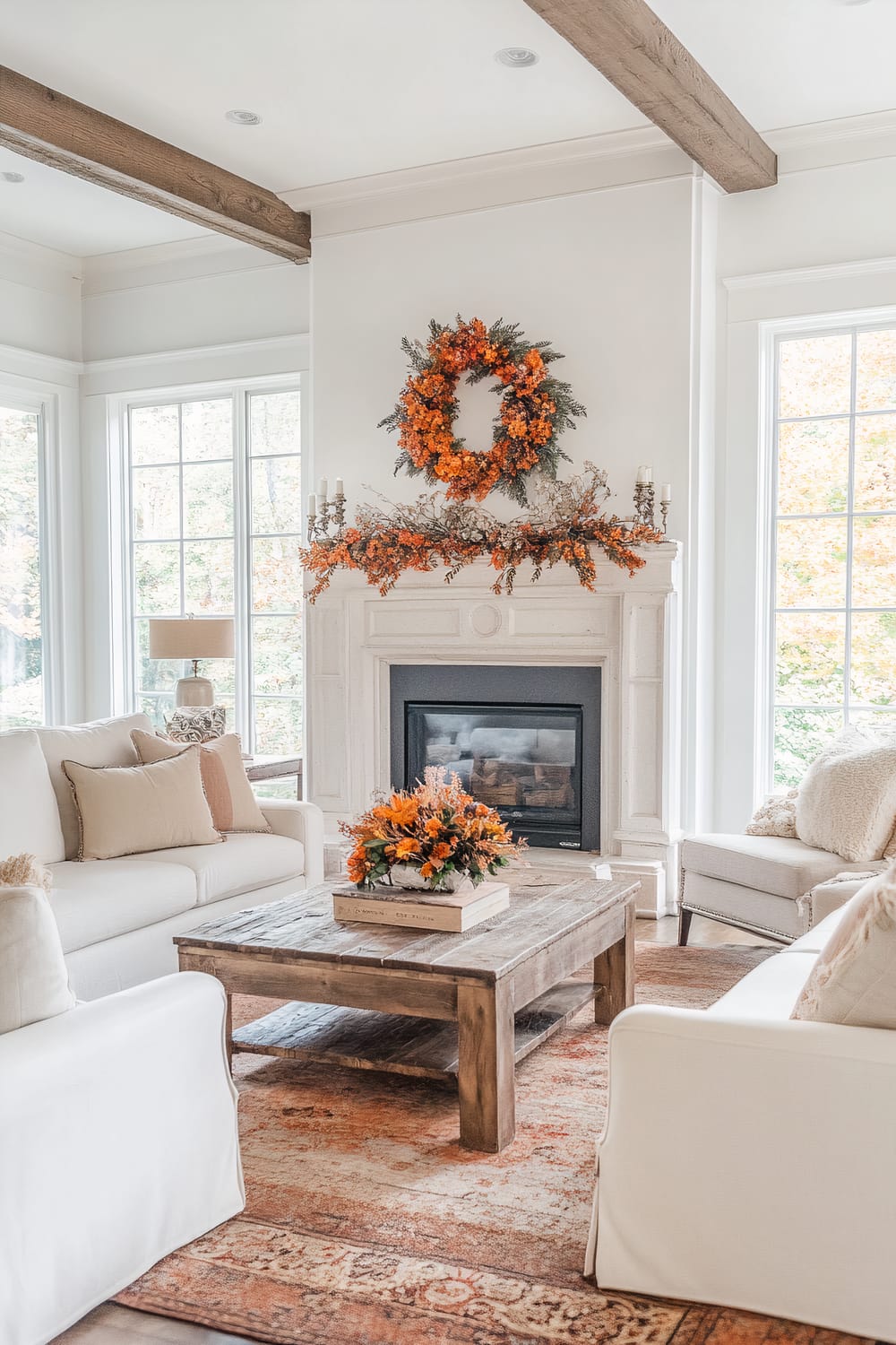 A bright, elegant living room featuring a central fireplace adorned with a vibrant orange autumn wreath and matching garlands. The room is furnished with white, upholstered sofas and a wooden coffee table decorated with an orange floral arrangement. Exposed wooden ceiling beams and large windows on either side of the fireplace allow ample natural light to flood the room.