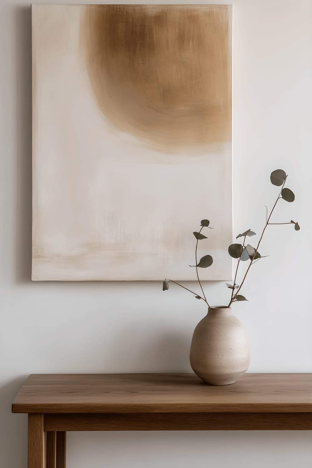 A minimalist Japandi-style console table made of smooth light oak wood stands against a blank wall. On the table, a handmade stoneware vase holds a single, delicate eucalyptus branch. Leaning against the wall above the table is a muted abstract painting emitting a calming aura. The scene is illuminated by soft, indirect lighting giving a tranquil vibe to the space.