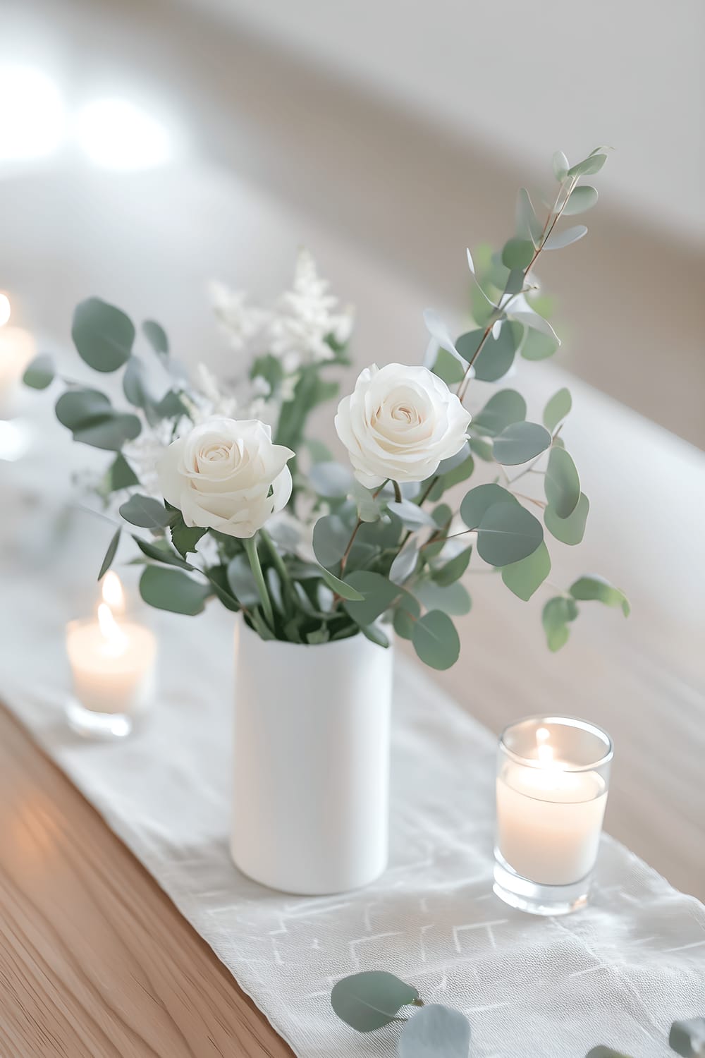 A centerpiece on a light oak table arranged in Scandinavian minimalist style featuring a white ceramic vase with a single eucalyptus sprig and some white roses, a subtle geometric runner, and a few small white candles housed in clear glass holders. Natural light diffuses softly across the arrangement, creating a serene, airy ambiance.