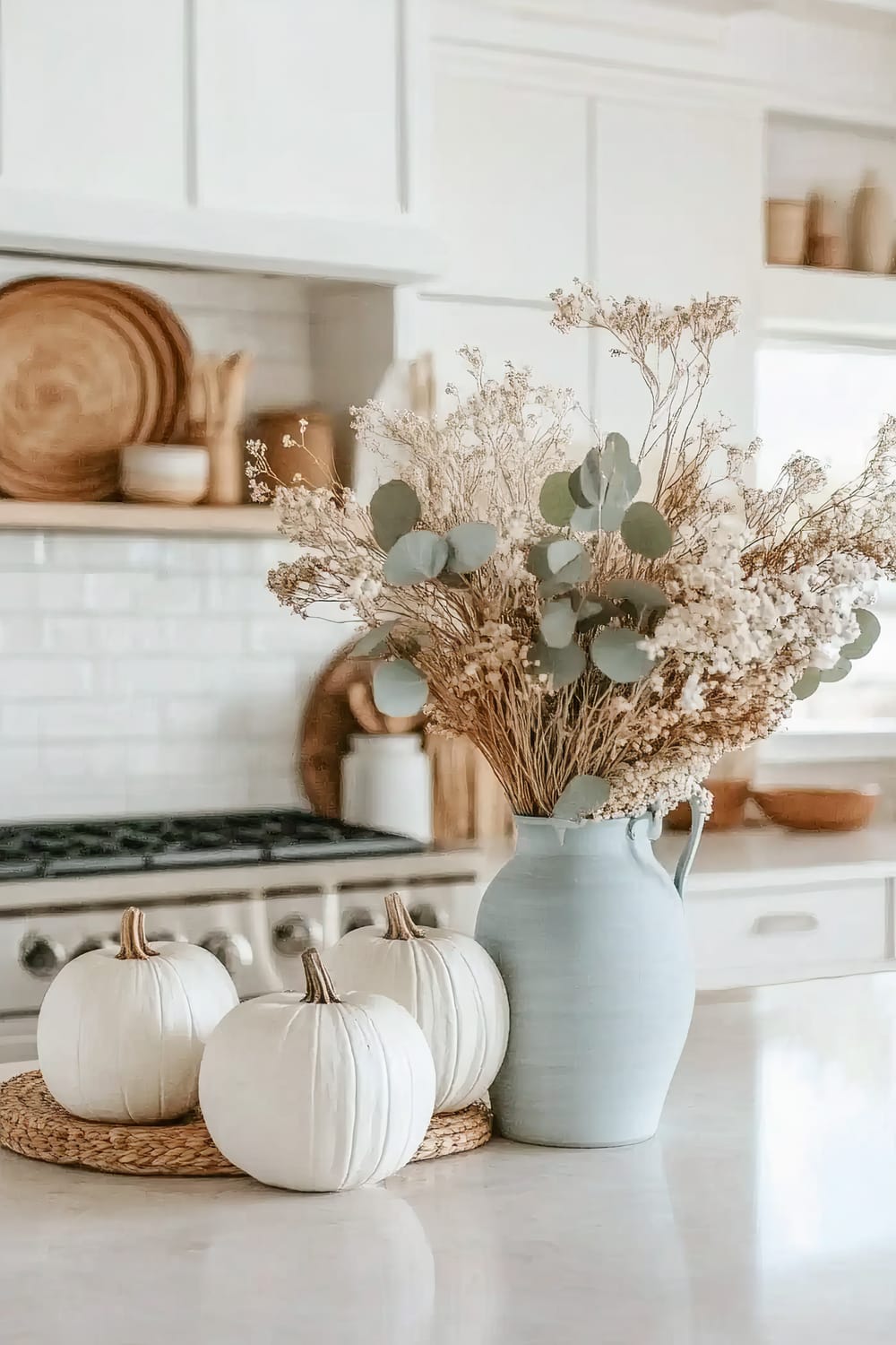 An aesthetically pleasing kitchen interior featuring white cabinetry, a white tiled backsplash, and a stovetop. In the foreground, there is a light blue ceramic vase filled with dried eucalyptus and baby's breath, placed next to three white decorative pumpkins on a woven mat. Natural wicker items are displayed on the open shelving in the background.