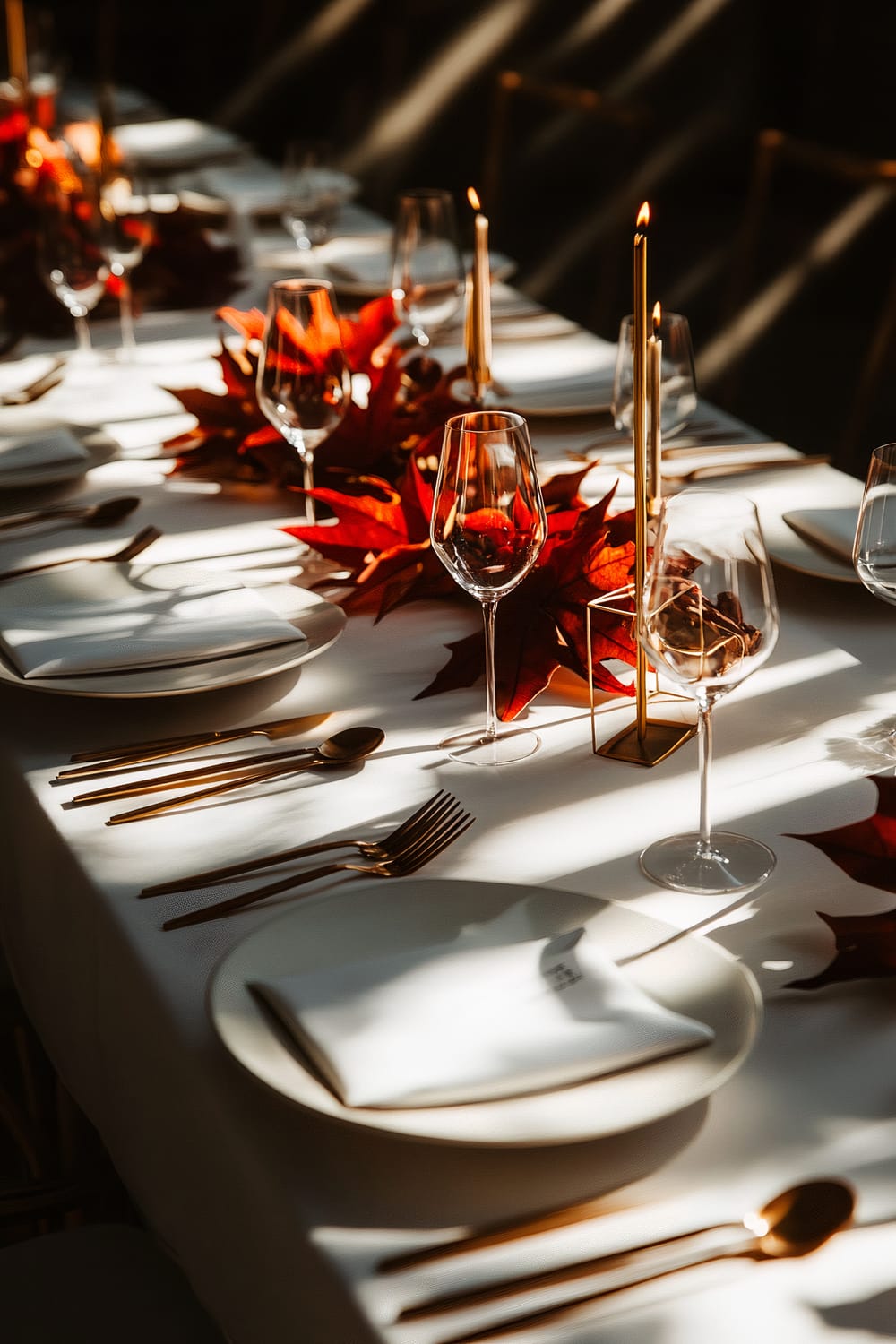 A contemporary Thanksgiving table setting with rich burgundy and gold accents. The table features white plates with gold flatware and wine glasses. A minimalist centerpiece consists of geometric candle holders and autumn leaves. Dramatic lighting casts elegant shadows, creating a sophisticated atmosphere.