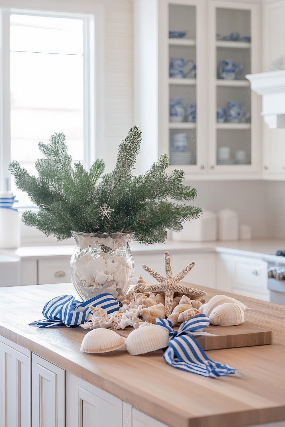 A coastal-style kitchen featuring white shaker cabinets and light wood floors. The kitchen is decorated with blue and white nautical-themed Christmas accents, including a large glass vase filled with white seashells, blue starfish, and evergreen branches. Blue and white striped ribbons, small driftwood ornaments, and potted seasonal plants enhance the festive atmosphere. Natural sunlight streams through large windows, creating a bright and airy ambiance.