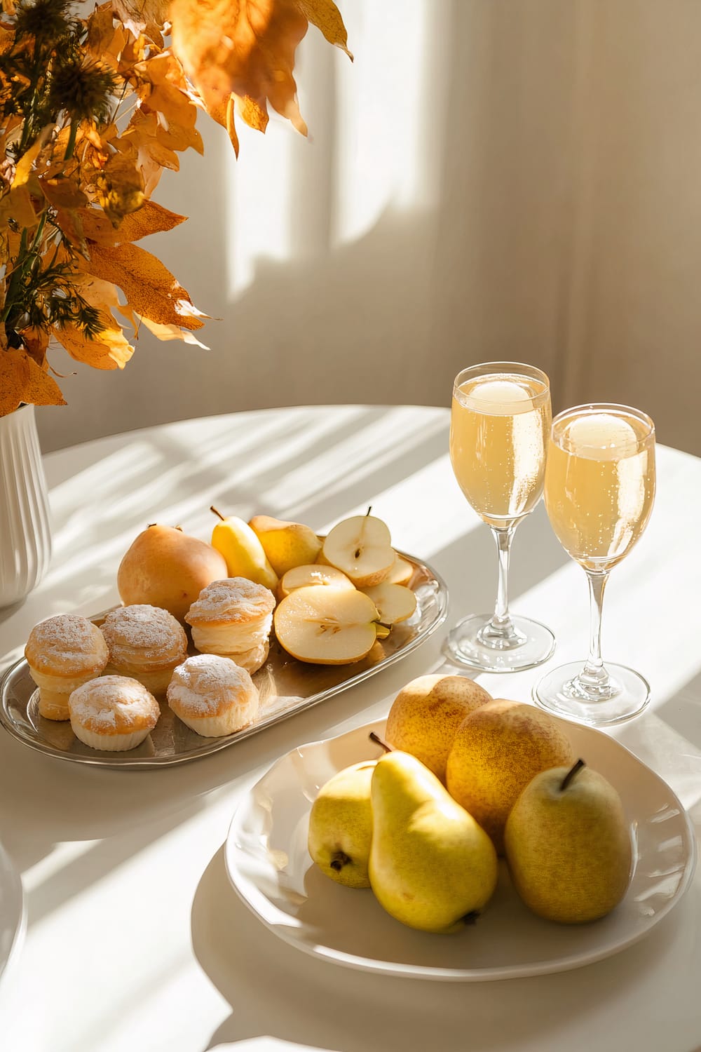 A minimalist table setting for a Friendsgiving brunch featuring a white ceramic platter with assorted pastries, a metal platter with sliced pears, and two elegant vintage glasses of mimosa. The sleek white table is softly lit, with autumnal foliage in a white vase enhancing the warm, inviting atmosphere.