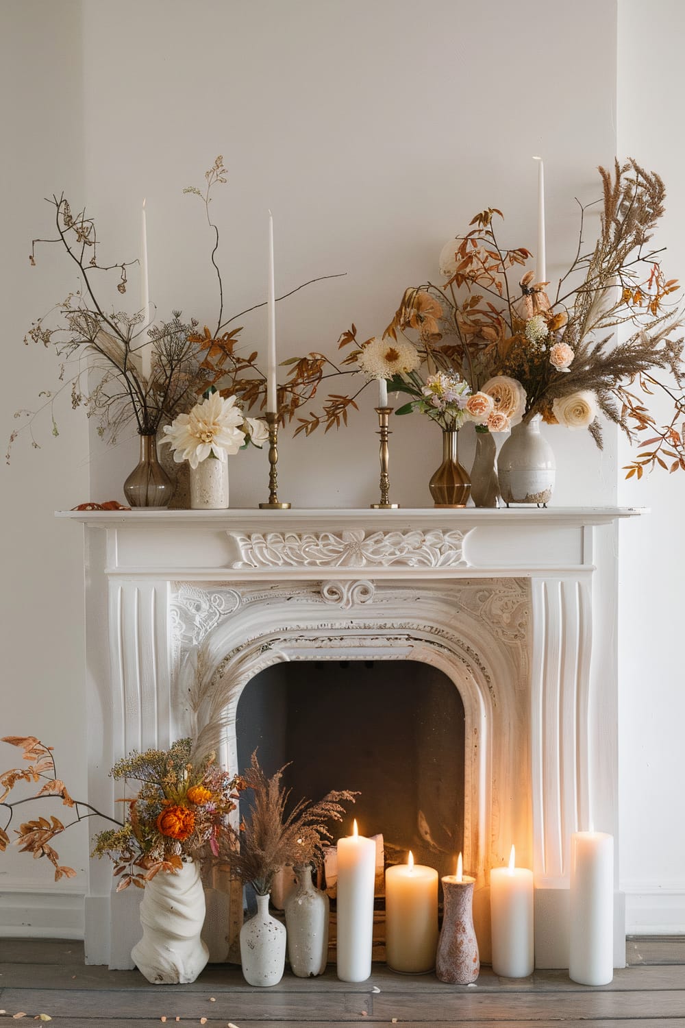 An ornate white fireplace mantel decorated with a collection of candles, flowers, and decorative branches. On top of the mantel, there are tall candles in gold holders, white and beige flowers in vases, and sprigs of autumnal foliage. At the base of the fireplace, there are more candles of different heights, a white and a reddish-brown vase with dried flowers and foliage.