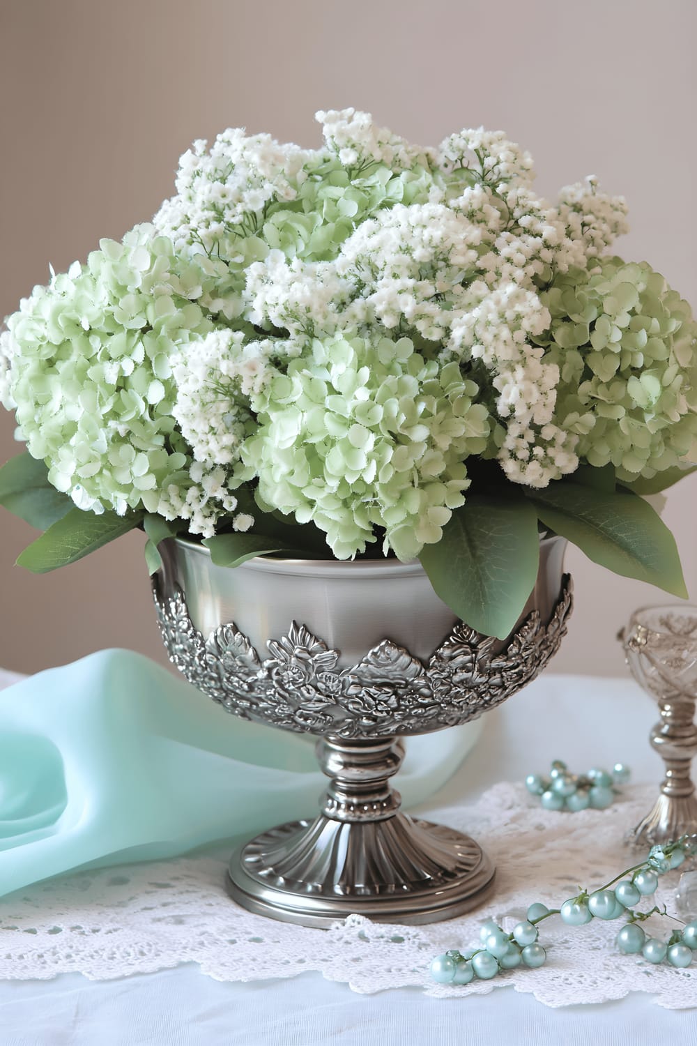 A centerpiece on a light mint tablecloth featuring a pewter bowl that is filled with fresh mint green hydrangeas and delicate white baby’s breath. The arrangement is accented with two pewter candle holders, scattered mint green glass beads, and small sprigs of greenery. Soft, natural lighting makes the cooler colors pop and brings a soothing, elegant feel to this table arrangement.