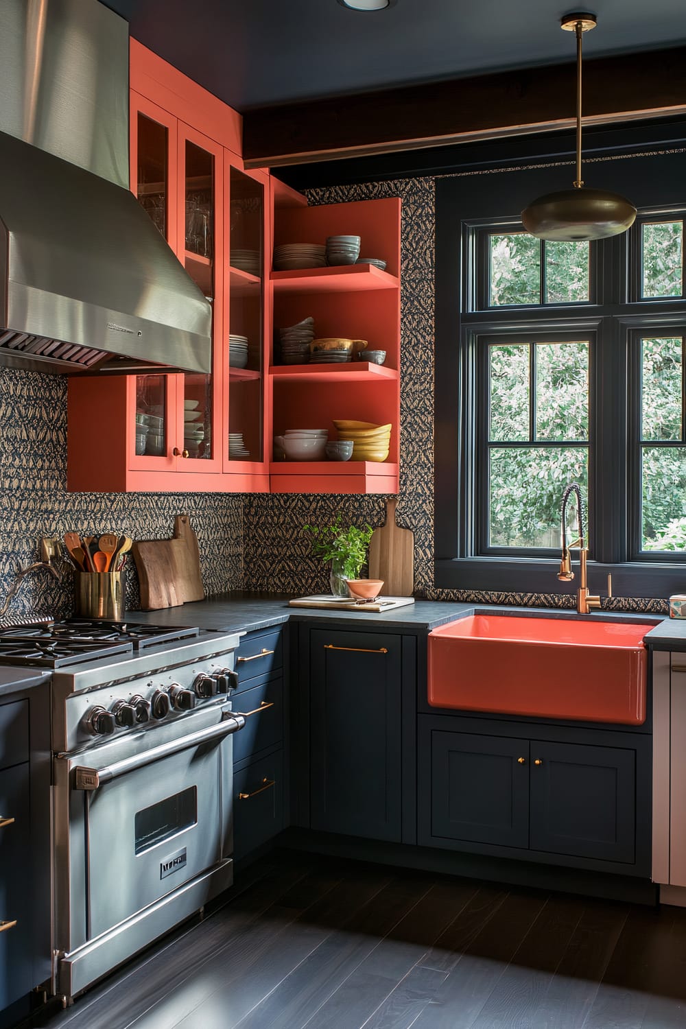 A sophisticated farmhouse kitchen features navy blue lower cabinets and bright coral upper cabinets creating vibrant color contrasts. The kitchen is equipped with a stainless steel range, built-in oven, and refrigerator. A large farmhouse sink with vintage fixtures is installed under a window, bringing in natural light. The dark slate floors and textured backsplash add depth, while colorful ceramic bowls are displayed on open shelves.