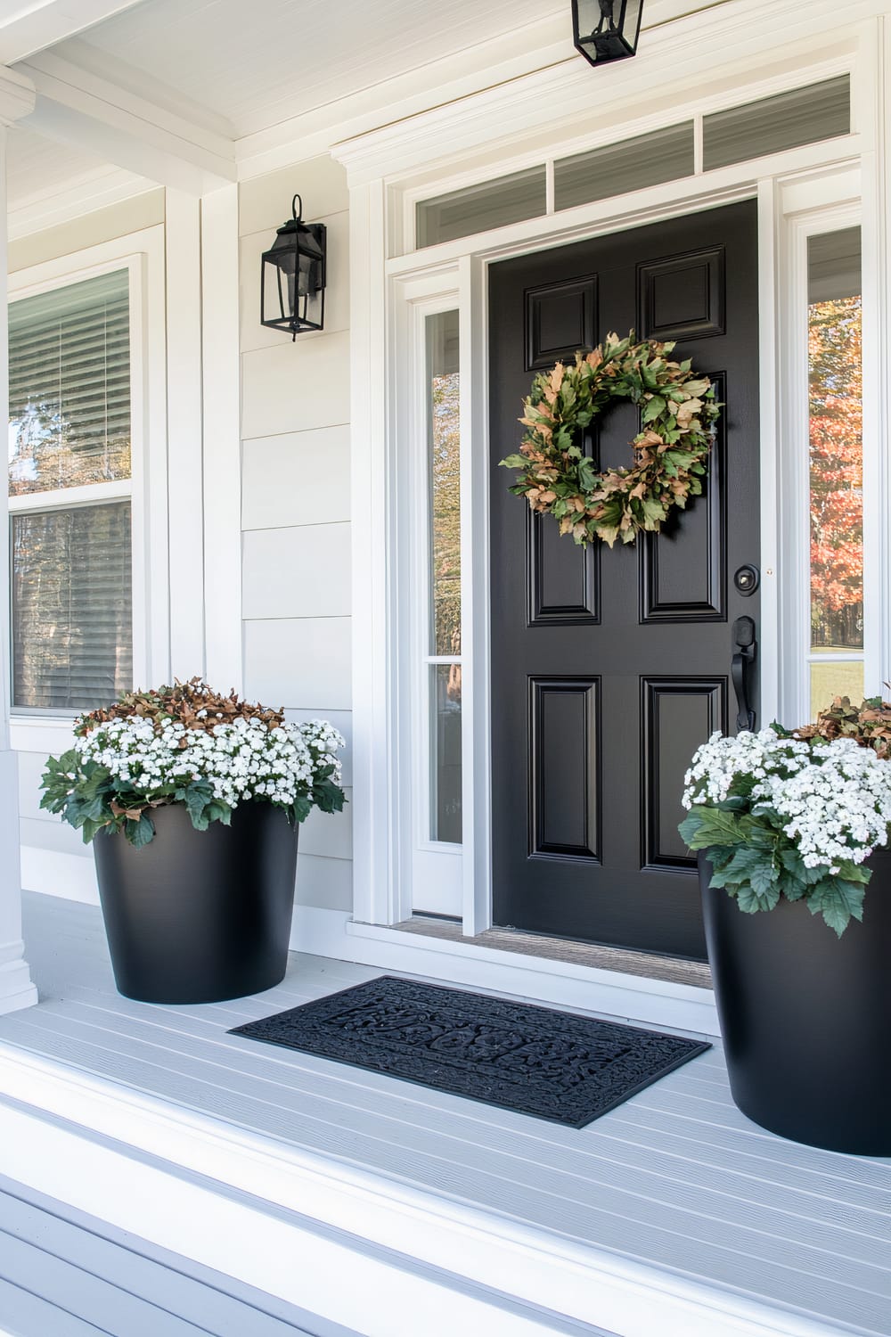 A front entrance featuring a black door with a decorative green and brown wreath. Flanking the door are two large black planters containing white flowers and green foliage. The porch is painted white with light gray decking and features wall-mounted black lantern lights on either side of the door. A black welcome mat lies in front of the door.