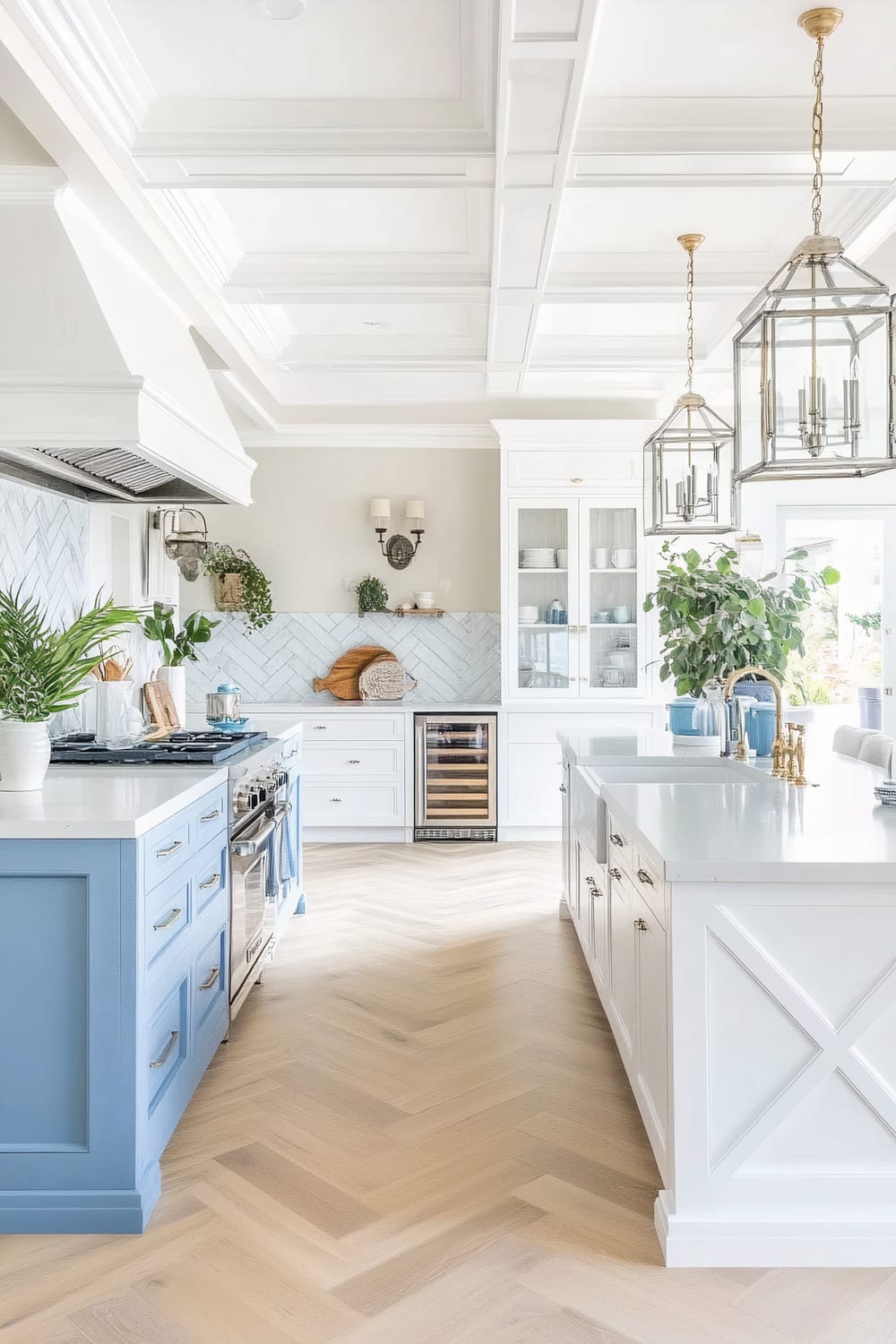 A bright and airy kitchen featuring blue and white cabinetry. The kitchen has a central island with golden faucets and white countertops. The walls are adorned with a herringbone tile backsplash, and there are various potted plants adding a touch of greenery. The floor has a light wood herringbone pattern, and large, geometric pendant lights hang over the island. The ceiling has an intricate coffered design.