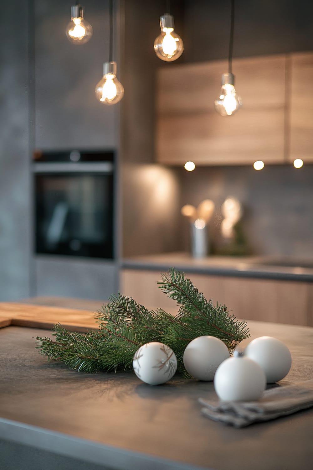 A Scandinavian-style kitchen countertop features minimalist Christmas decorations. It includes white ceramic ornaments and a single evergreen sprig with subtle decorations, under soft ambient lighting. The decor is enhanced by natural wood elements and sleek silver accents, creating an uncluttered and elegant space.