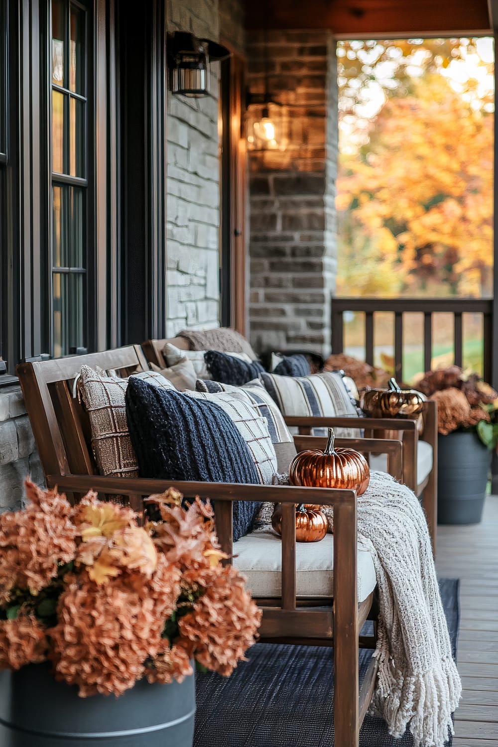 A front porch is adorned with autumn-themed decorations and plush seating. Wooden benches are accessorized with a mix of blue and neutrally-toned textured pillows, and cozy throws. Small metallic pumpkins are placed on the benches, enhancing the fall vibe. Large potted plants with burnt orange and brown foliage complement the seasonal decor. The background includes a stone exterior wall, black-framed windows, and an outdoor lantern-style light fixture. An array of autumnal trees with yellow and orange leaves can be seen beyond the wooden railing.