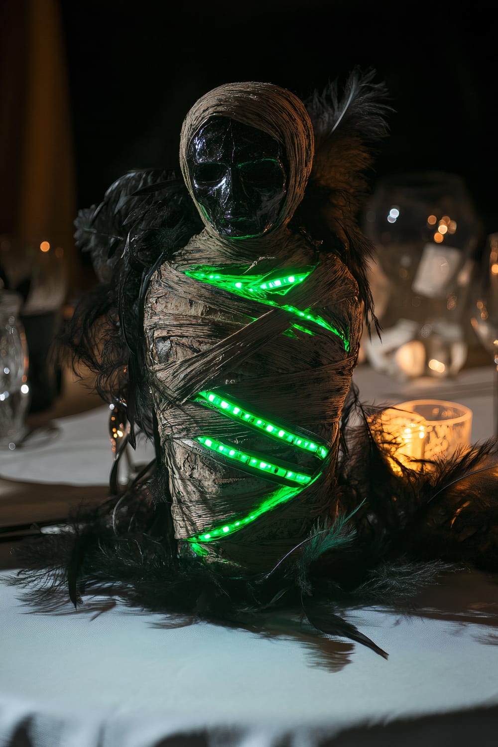 An eerie mummy centerpiece is placed on a simple white tablecloth. The mummy appears ancient, wrapped in tattered bandages with glowing green LED strips running through them. It is surrounded by dark feathers and a cracked mirror that reflects the green light. There is atmospheric, dim lighting with some blurred, warm lights in the background.