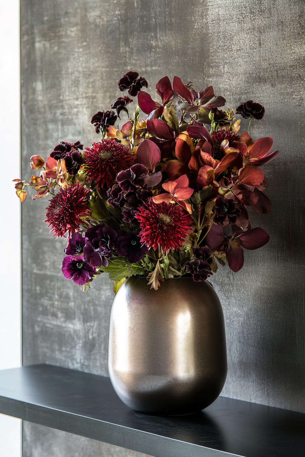 A metallic vase holding a vibrant arrangement of deep red and purple flowers sits on a dark shelf. The background is a textured gray, creating a contrast with the colorful flowers.