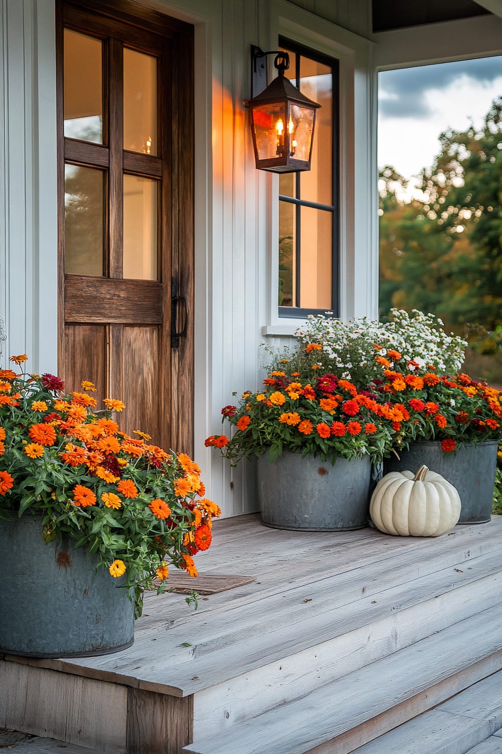 A front porch with wooden steps and a door, decorated with large pots of vibrant orange and red flowers as well as a white pumpkin. A warm wall-mounted lantern beside the door illuminates the entrance.