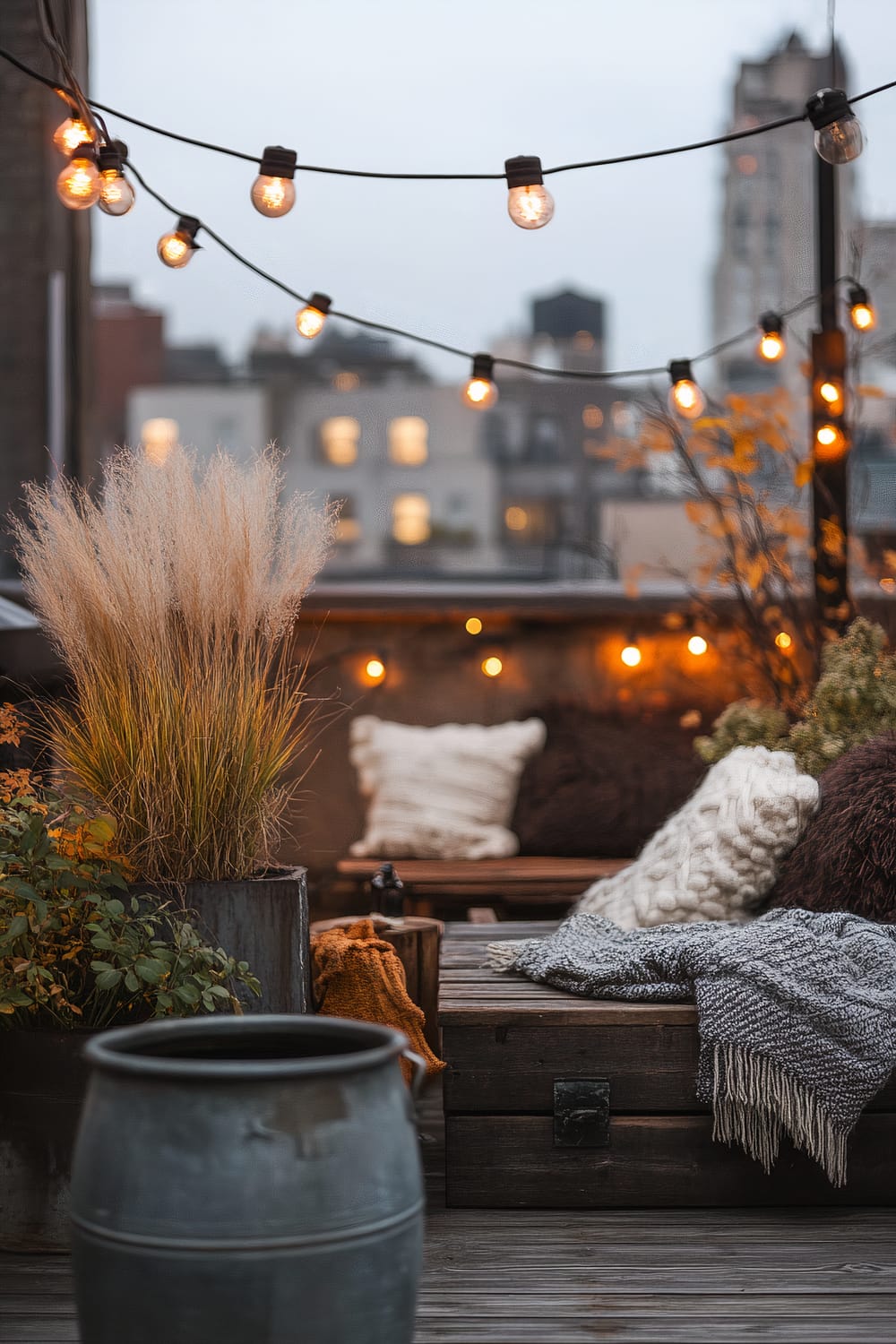 A cozy outdoor rooftop patio at dusk, illuminated by hanging bulb string lights. The space is adorned with various plants in pots and features a rustic wooden bench draped with blankets and cushions. The soft glow of the lights highlights the texture of the fluffy cushions and blankets, creating a warm, inviting atmosphere. The background reveals city buildings slightly blurred, adding a sense of urban charm.