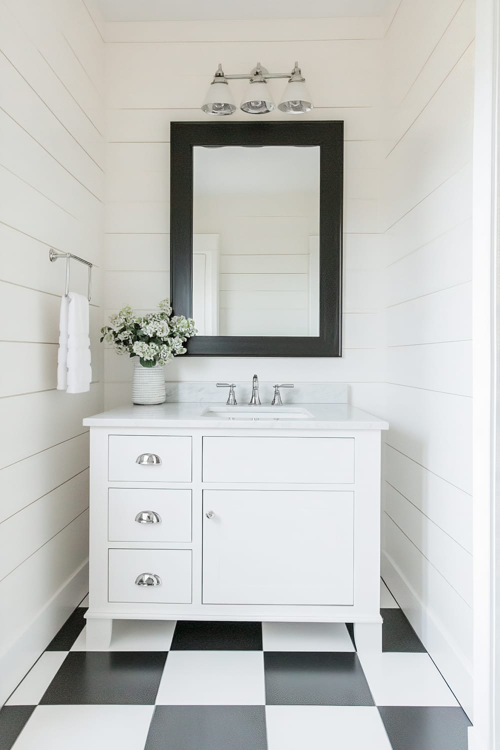 This image shows a modern bathroom with a monochromatic color palette. The walls are lined with horizontal white shiplap, providing a clean and bright backdrop. The focal point is a white vanity with a marble countertop and chrome fixtures. Above the vanity hangs a substantial black-framed mirror, topped with a trio of chrome light fixtures. A vase of small white flowers sits on the counter next to the sink. The floor features a bold black and white checkered pattern that adds a dramatic contrast to the space.