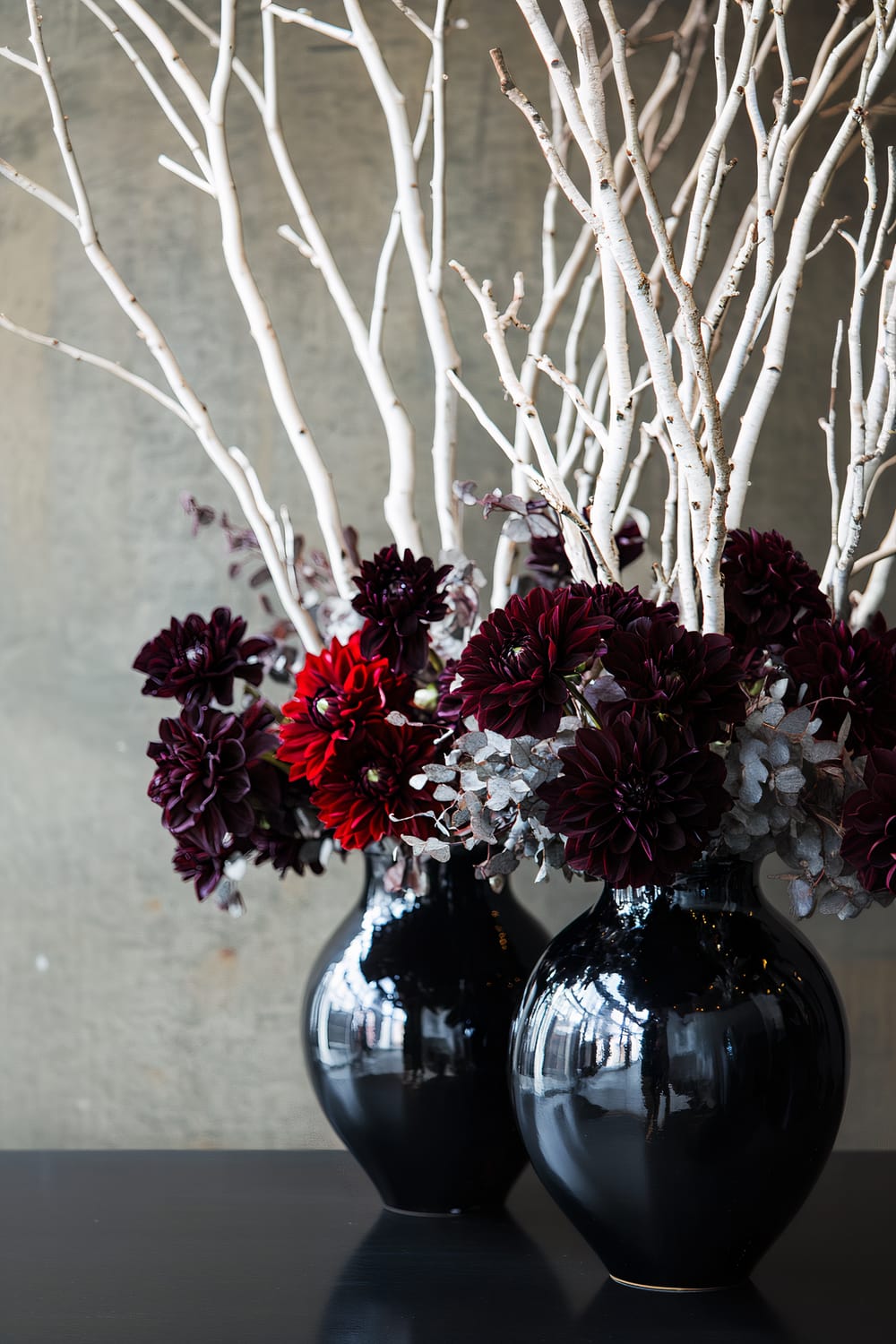 A pair of black ceramic vases filled with dark red dahlias, white painted branches, and silver eucalyptus leaves, set against a neutral gray background.