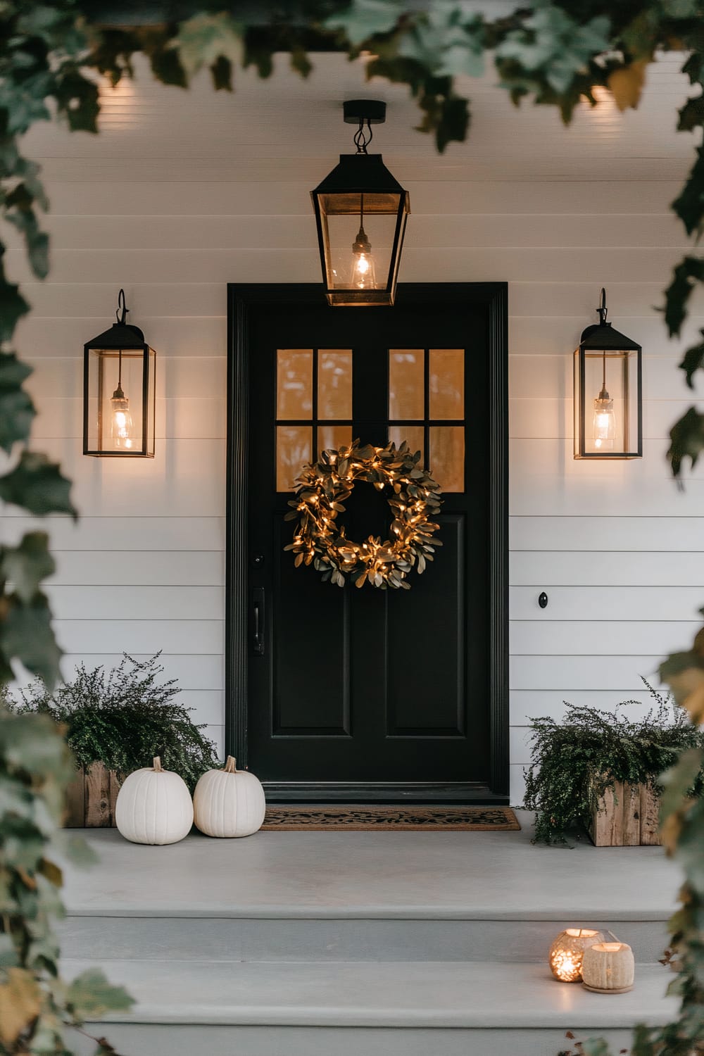 A porch scene with a black front door adorned with a gold wreath, flanked by two black frame lanterns with lit bulbs. A larger lantern hangs above the door. Two white pumpkins and potted greenery are placed by the door. Small candle holders are on the steps, and leafy branches frame the image.