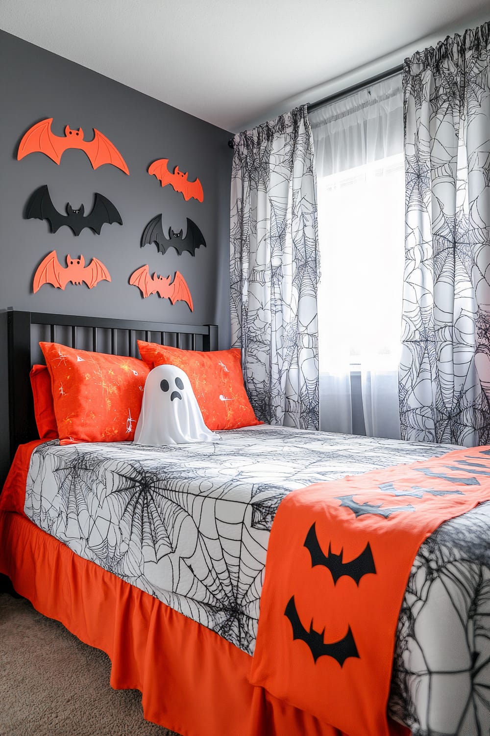 A Halloween-themed bedroom with a twin bed featuring an orange skirt, spiderweb-printed sheets, and matching pillowcases. A small, friendly-looking ghost decoration sits on the bed. The room's gray wall is adorned with orange and black bat-shaped wall decorations. The window is covered with white curtains also printed with spiderweb patterns, allowing natural light into the room.