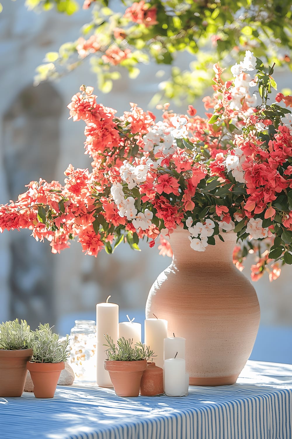 A large ceramic urn filled with lush bougainvillea and olive branches, surrounded by white candles and small terracotta pots, rests on a blue and white striped tablecloth. The scene is bathed in bright, natural sunlight, giving it an overall vibrant and ripe ambiance reminiscent of a Mediterranean coastal home.