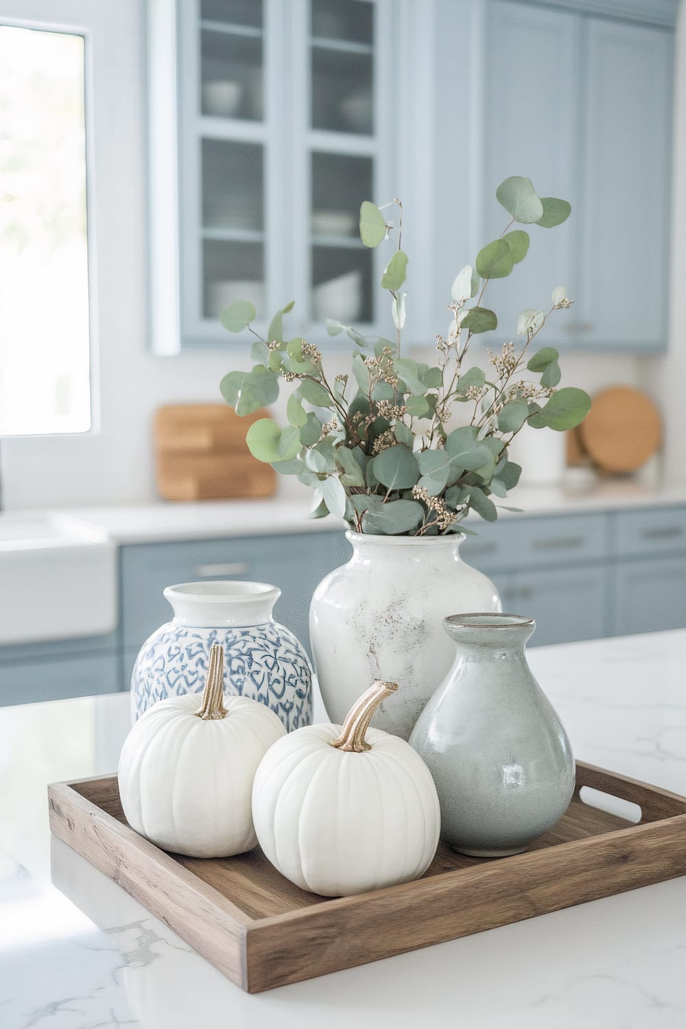 A serene kitchen setting featuring a wooden tray on a white marble countertop. The tray holds two white pumpkins and three ceramic vases, one of which contains eucalyptus branches. Blue-gray cabinetry with glass doors is visible in the background, along with wooden cutting boards, adding a rustic yet modern touch to the space.