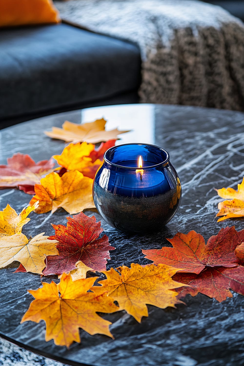 An elegant coffee table with a dark marble surface is decorated with fall leaves in yellow and red hues. At the center of the table, there is a cobalt blue candle holder with a lit candle inside, creating a sharp contrast with the autumn colors of the leaves. In the background, part of a grey sofa with a textured blanket and an orange pillow is visible.