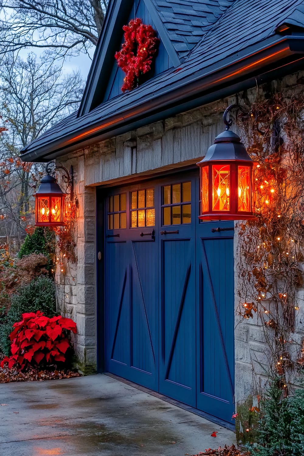 A garage door painted in steel blue with red lanterns and a red wreath hanging above. Two red lanterns, one on each side of the door, illuminate the entrance, casting a warm glow. Festive decorations including string lights and red poinsettias are placed near the door. The surroundings feature stone walls and some greenery.