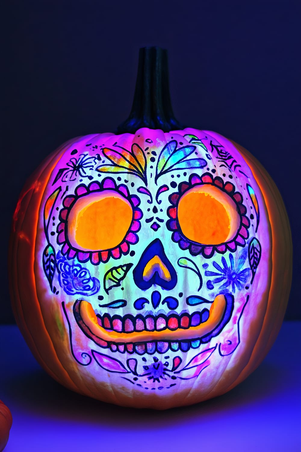 A carved and painted pumpkin resembling a colorful sugar skull, illuminated with vibrant neon colors under blacklight, highlighting intricate Day of the Dead motifs and patterns.