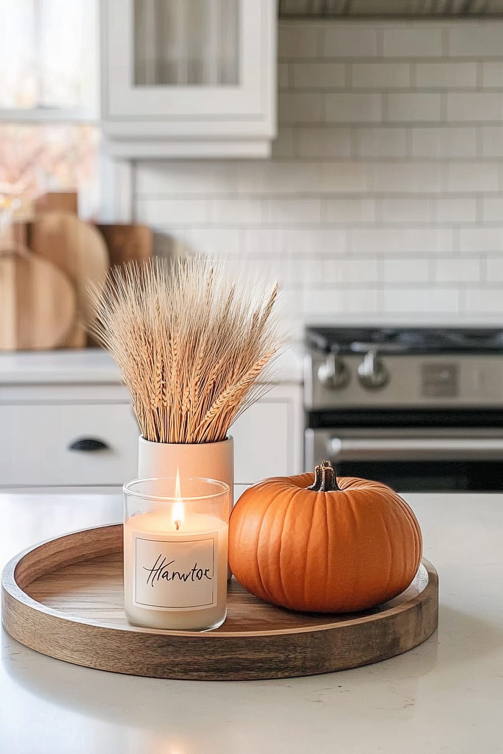 A kitchen countertop features a wooden tray that holds a lit candle, a small pumpkin, and a bundle of wheat stalks in a white vase. The background includes white cabinetry with black handles, a stove, and a tile backsplash.