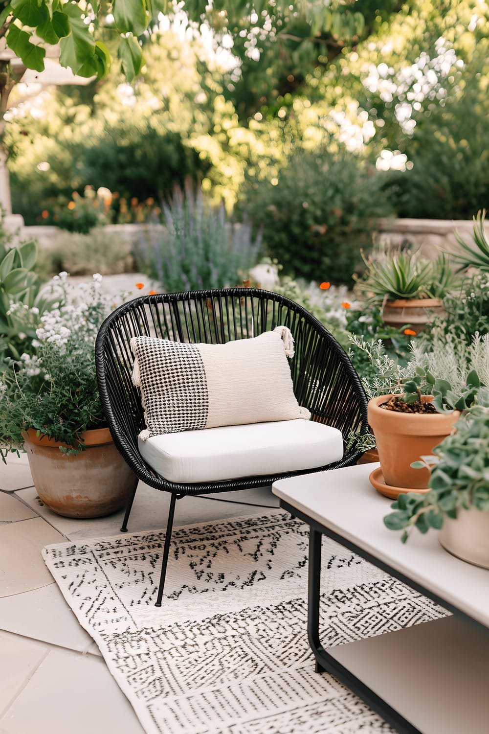 The image portrays a minimalist modern outdoor space with a sleek black rattan chair placed on a neutral outdoor rug. The chair is accessorized with neutral-toned cushions. Also on the rug is a low-profile coffee table housing a modern planter that serves as a centerpiece. The decor of the area includes a carefully selected array of plants in monochrome and terracotta pots.