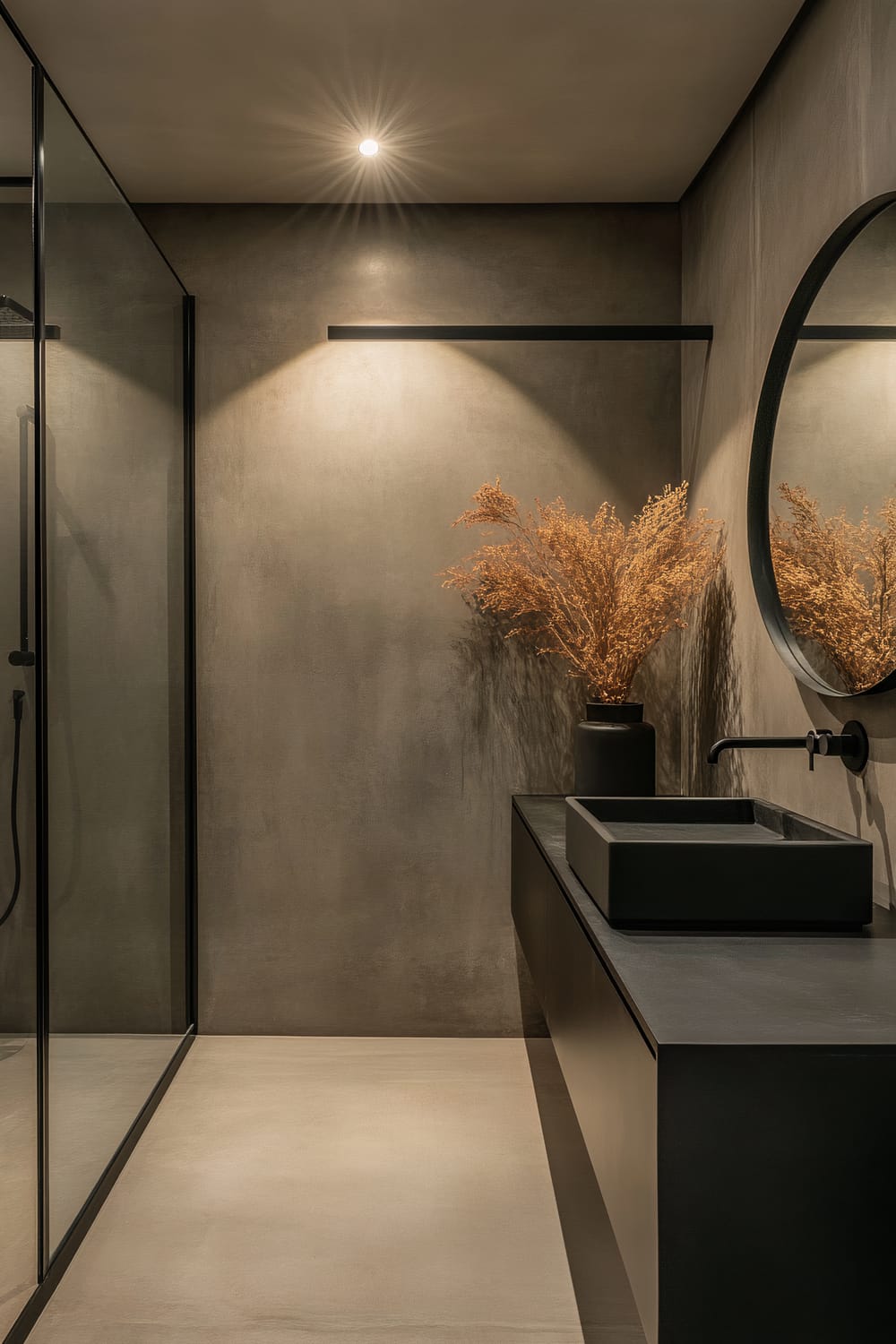 An elegant and minimalist bathroom featuring a black floating vanity with a square black vessel sink and a wall-mounted black faucet. Adjacent is a large circular mirror, reflecting the neutral tones of the space. A single spotlight on the ceiling illuminates the room, casting a warm glow on the taupe walls and polished concrete floor. A glass-enclosed shower is visible on the left, and a vase with dried plants adds a touch of organic texture.