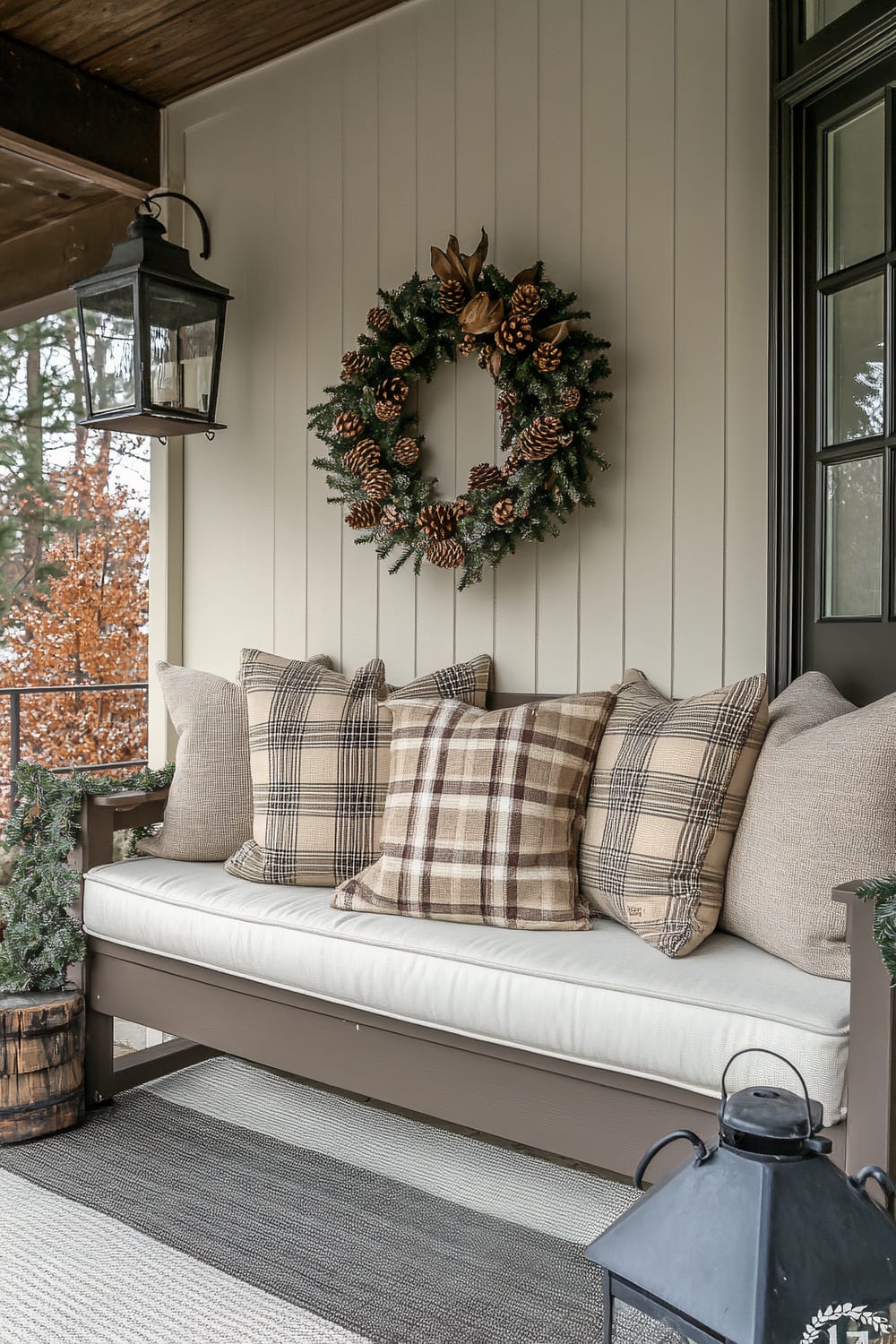 An outdoor seating area featuring a beige cushioned bench adorned with plaid and solid tan pillows. Above, a decorative Christmas wreath made of pinecones and greenery hangs on the beige paneled wall. To the left, a vintage-style black lantern is mounted on the wall. The bench sits on a gray and white striped rug, with a black lantern placed on the floor nearby.