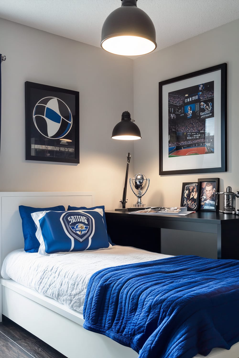 A bedroom with a sporty theme featuring a white futon bed and a blue sports-themed blanket. The walls are light grey, and the space is illuminated by a single overhead lamp. A black minimalist desk stands next to the bed, displaying a single sports trophy and a few photos. Two sports-themed posters are mounted on the wall above the bed and desk.