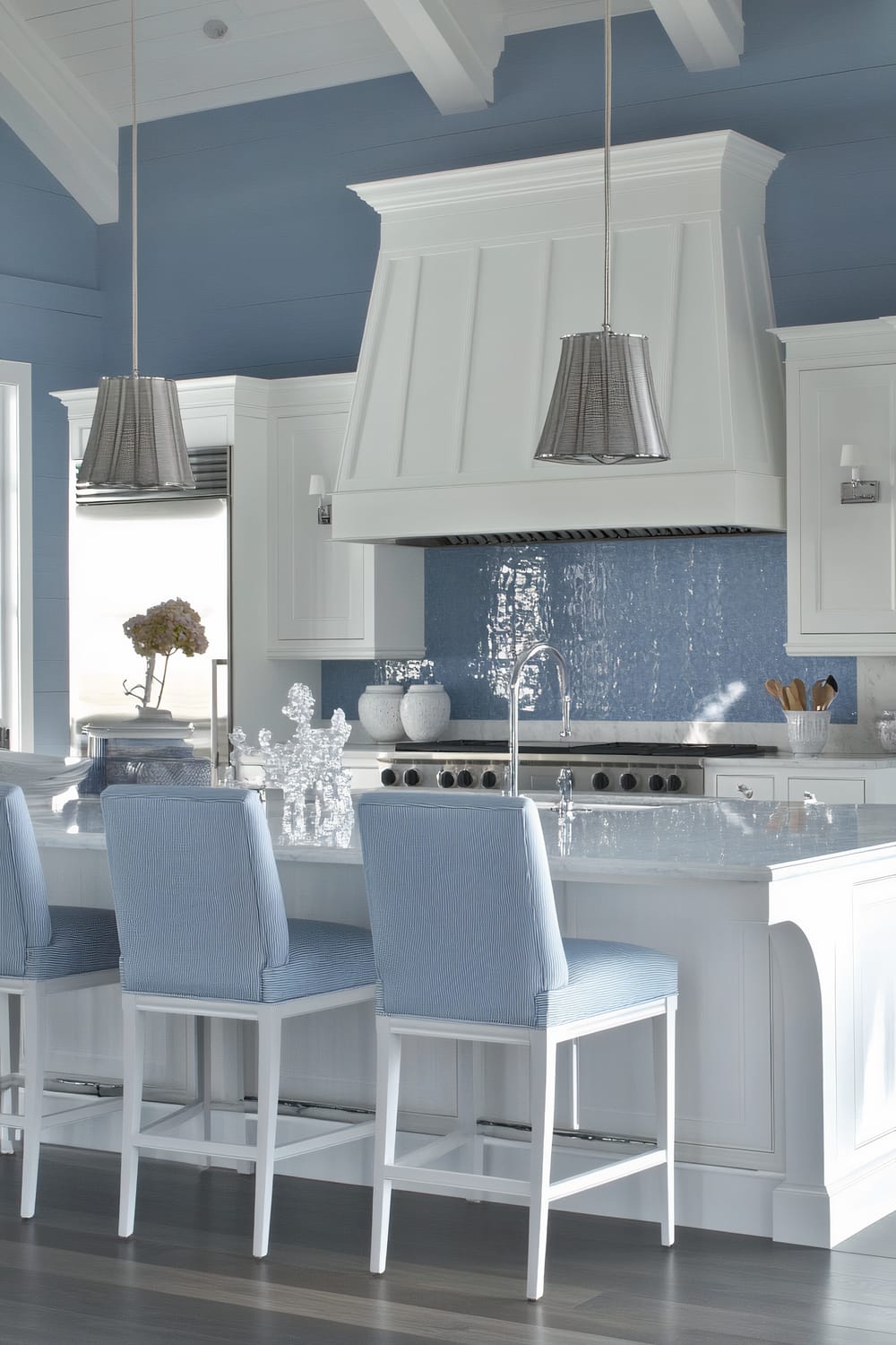 A sophisticated kitchen featuring a light blue and white color scheme. The kitchen island has white cabinetry with a marble countertop, adorned with crystal decorative pieces. Three light blue upholstered bar stools with white wooden frames offer seating at the island. Above the island, there are two pendant lights with silver shades. The walls are painted in a pastel blue, contrasted by white cabinetry and a large white range hood over the stove. The backsplash is a shimmering blue tile that enhances the serene aesthetic of the space.