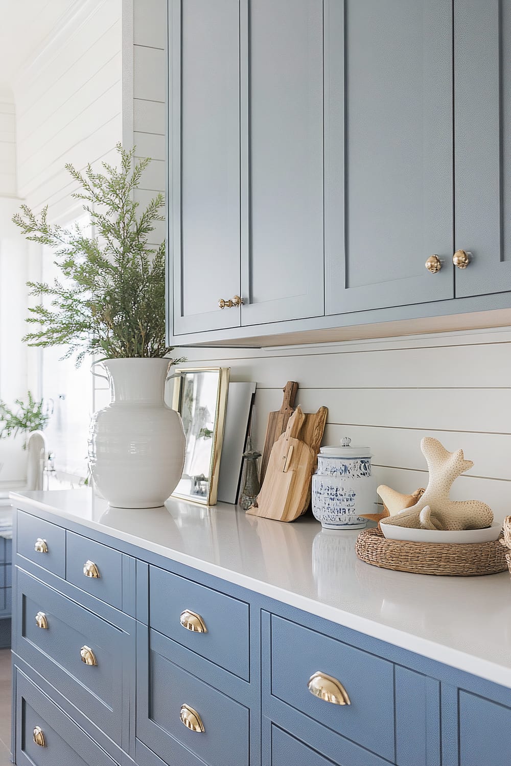 A stylish kitchen displaying a serene, coastal-inspired aesthetic. The cabinetry is painted in a muted blue-grey hue, accented by brass hardware knobs and handles. A pristine white countertop is adorned with a large white vase containing lush green foliage, wooden cutting boards, a blue and white ceramic jar, and decorative objects including a woven basket with starfish figures. The backdrop features horizontal shiplap paneling, also painted white, contributing to the bright and airy feel of the space.