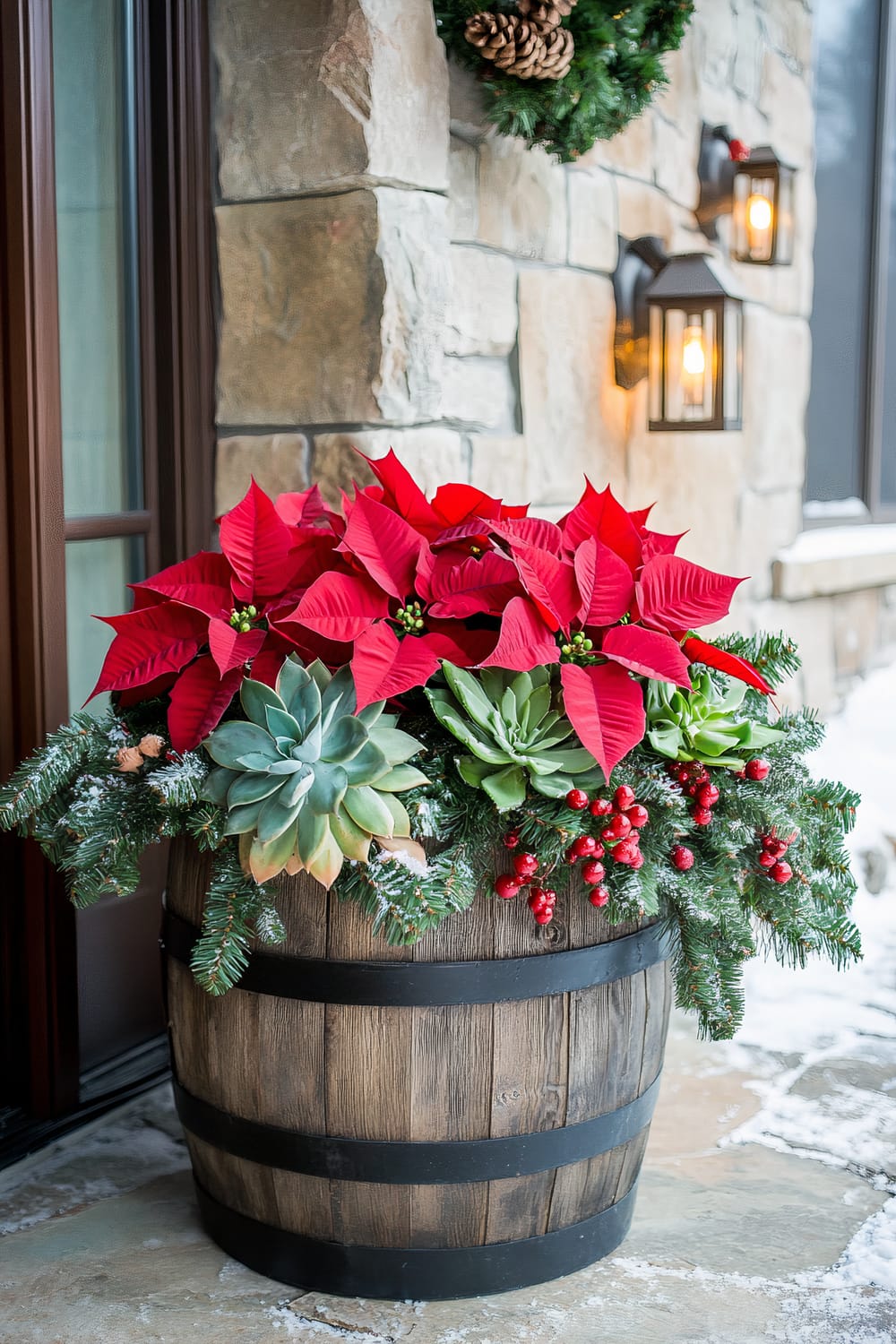 A rustic reclaimed wood barrel planter is filled with vibrant red Poinsettias, trailing green succulents like Echeveria, and glossy green Aloe Vera plants. The arrangement is adorned with burlap ribbons, small wooden ornaments, and illuminated by warm amber lantern lights. The planter is placed on a farmhouse-style stone porch with artificial frost resembling softly falling snow.